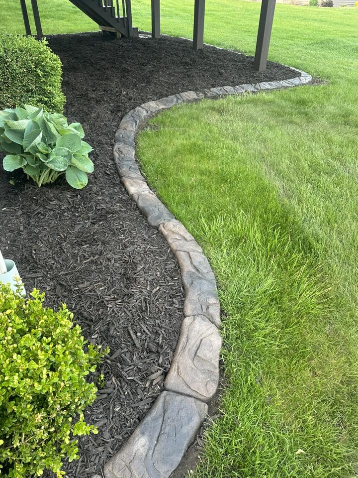 Curved stone edging separating a dark mulched garden bed from a green lawn beneath a deck, with shrubs and hostas nearby.