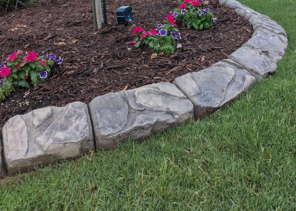 Stone edging surrounds a flower bed with mulch and bright pink and purple petunias, adjacent to green lawn.