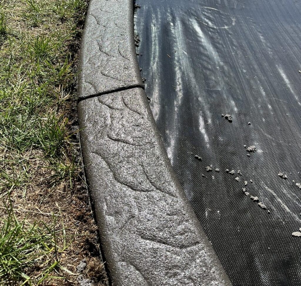 Cracked concrete edging separating a grassy lawn from a black landscape fabric sheet.